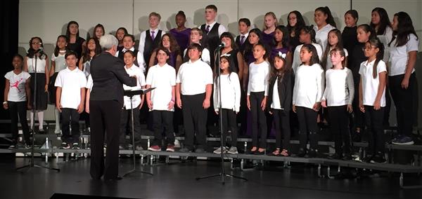 Several elementary students, dressed in black and white being conducted in a song by an adult female teacher